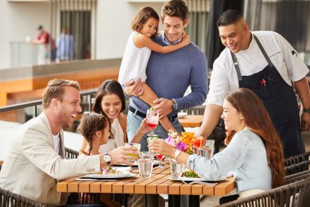 A imagem mostra uma família sentada à mesa, fazendo uma refeição no restaurante, enquanto o garçom está servindo os alimentos