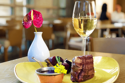 Peça de carne com beterrabas e recipiente de molho sobre prato amarelo. Ao lado, uma taça de vinho branco e decoração de flor vermelha em vaso branco.