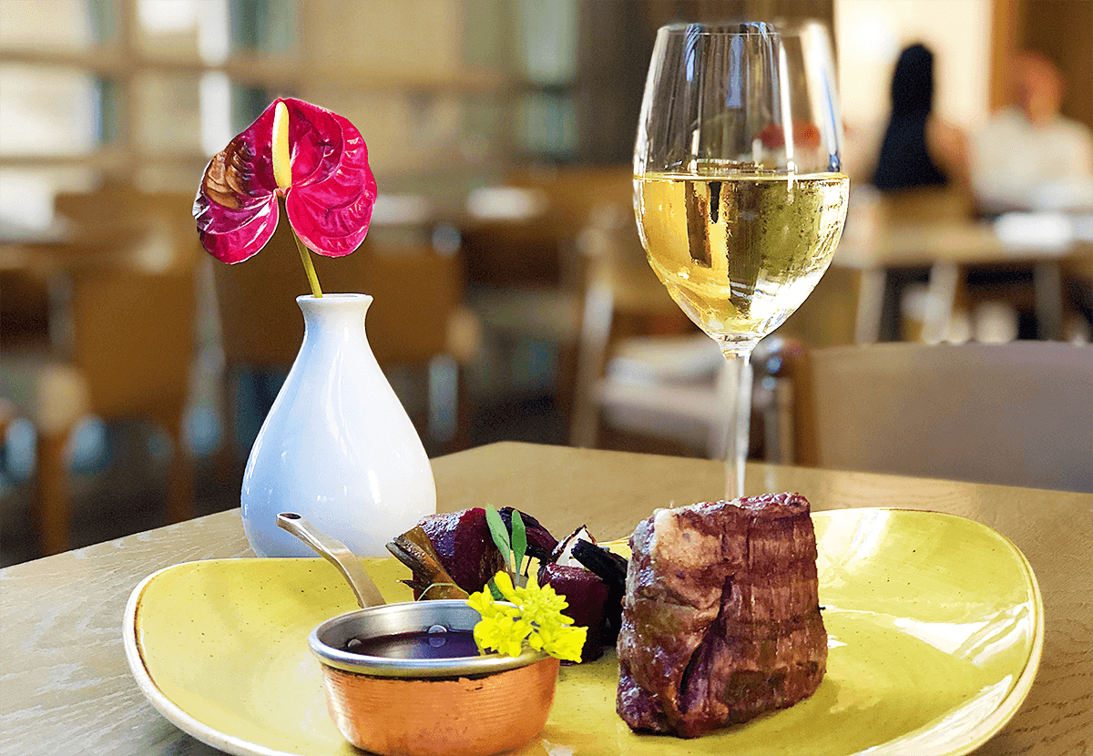 Peça de carne com beterrabas e recipiente de molho sobre prato amarelo. Ao lado, uma taça de vinho branco e decoração de flor vermelha em vaso branco.